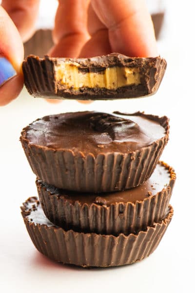 A hand holds a chocolate peanut butter cup with a bite taken out over a stack of other candies.