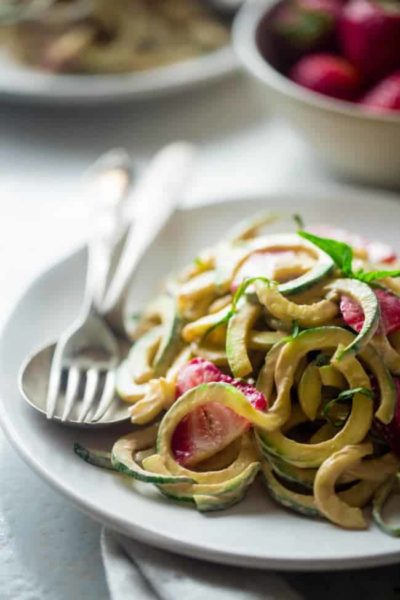 Zucchini noodles are on a plate with two forks. There are strawberries and a creamy sauce on the noodles.