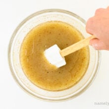 A hand holds a spatula and reaches in to stir golden colored liquid in a bowl.