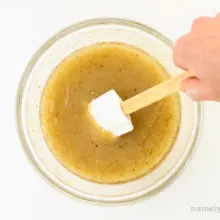 A hand holds a spatula and reaches in to stir golden colored liquid in a bowl.