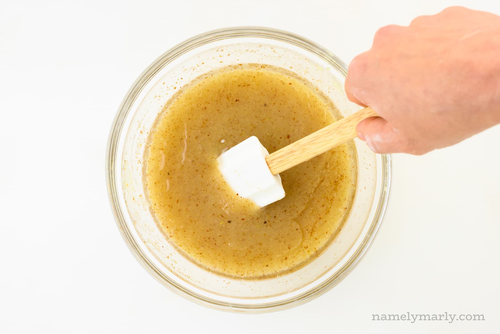 A hand holds a spatula and reaches in to stir golden colored liquid in a bowl.