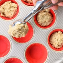 A hand holds a scoop, dropping batter into muffin compartments in a muffin pan.