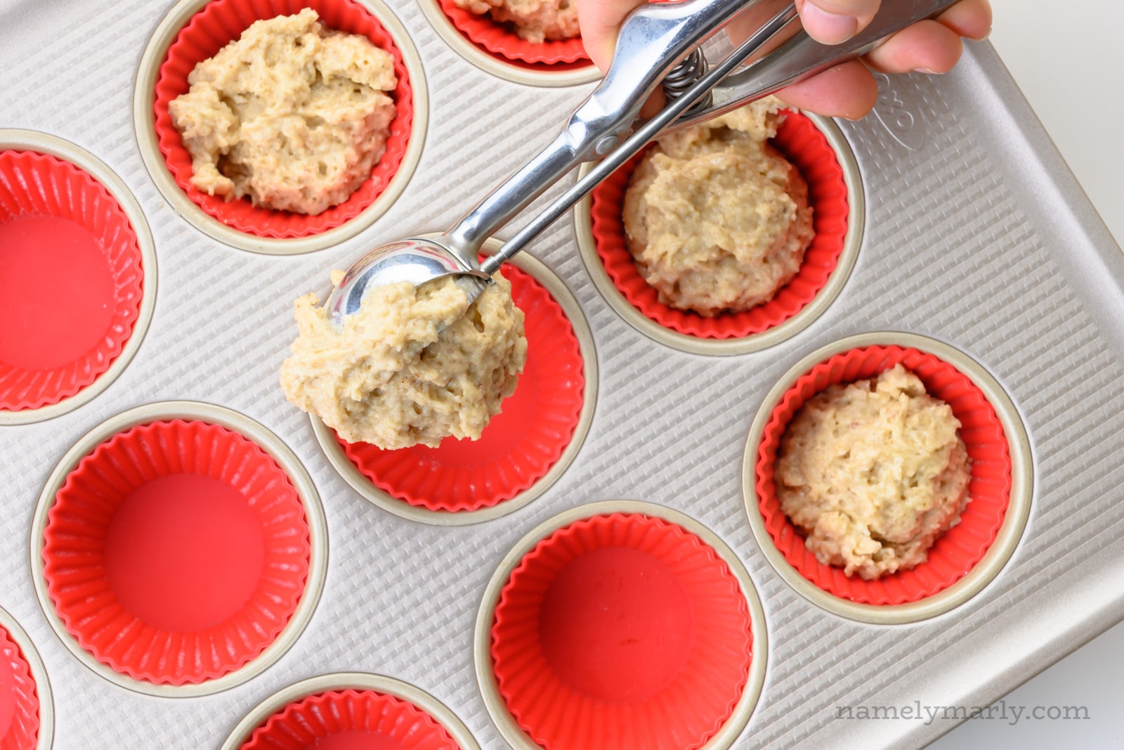 A hand holds a scoop, dropping batter into muffin compartments in a muffin pan.