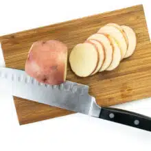 A red potato sits on a cutting board next to a butcher's knife. Several thin slices of potatoes sits next to it.