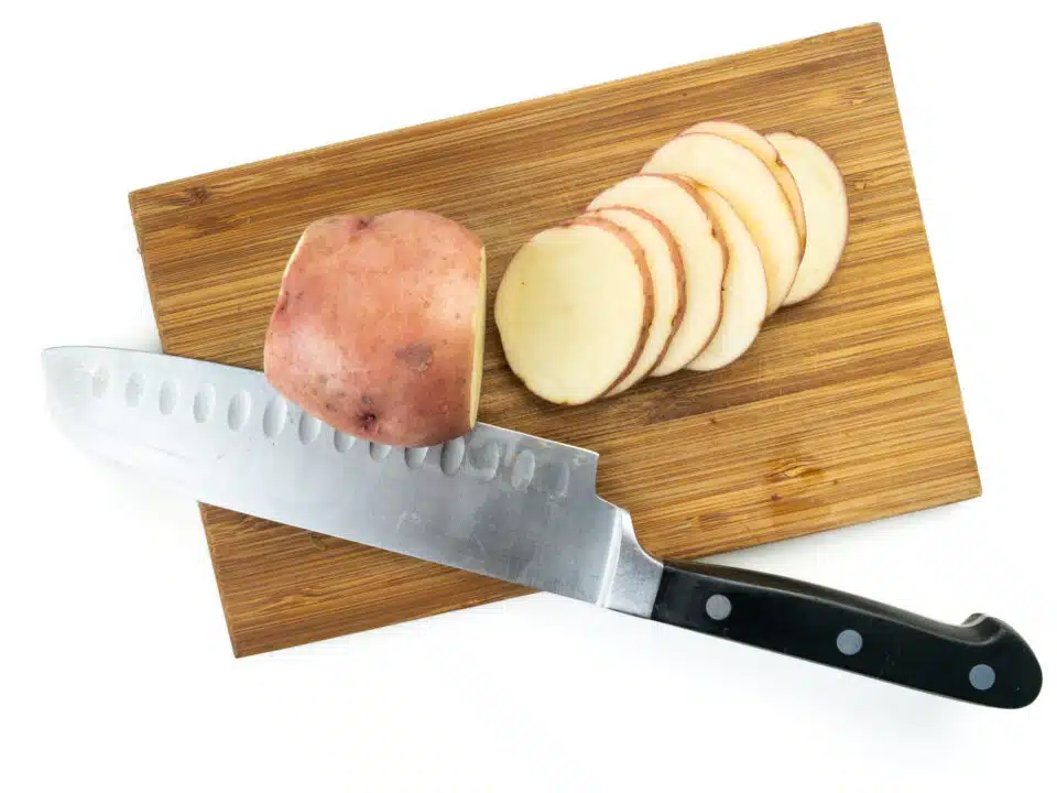 A red potato sits on a cutting board next to a butcher's knife. Several thin slices of potatoes sits next to it.