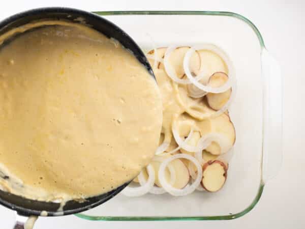 A pan of cream sauce is being poured over layers of potatoes and onions in a baking dish.