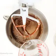 Hot water in a pyrex measuring cup is being poured into a mixing bowl of a stand mixer with chocolate cake batter in it.