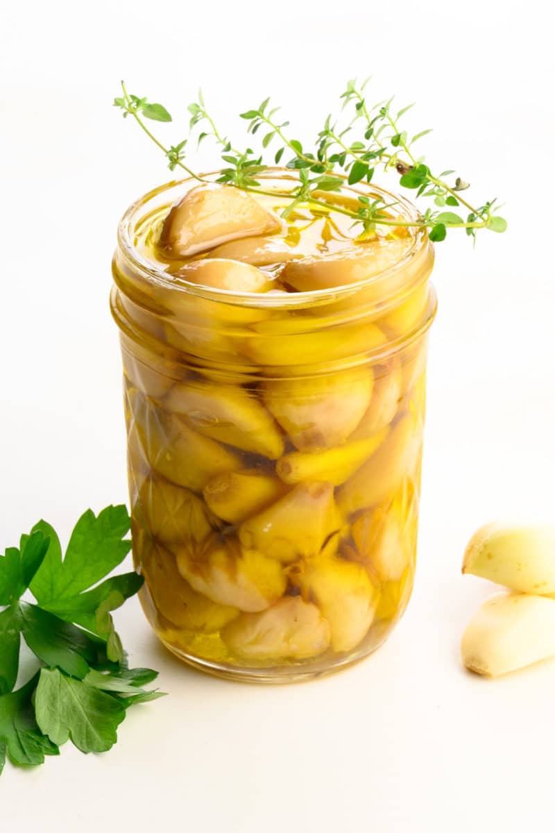 A mason jar holds many cloves of cooked garlic sitting in olive oil. There are cloves are garlic beside it along with green herbs.