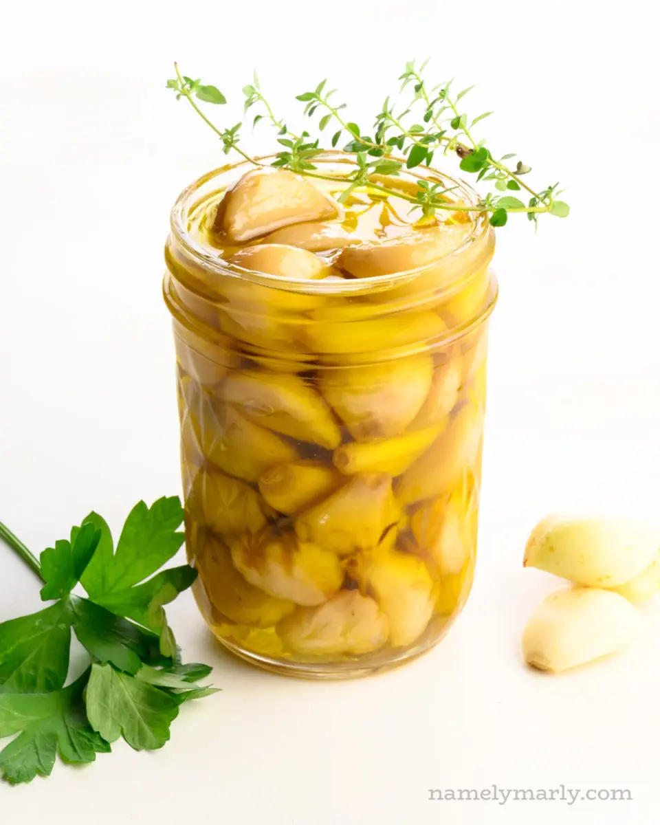 A mason jar holds many cloves of cooked garlic sitting in olive oil. There are cloves are garlic beside it along with green herbs.