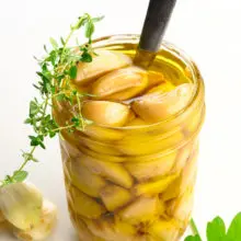 A mason jar holds garlic confit with a spoon. There are green herbs on top of and beside the jar.