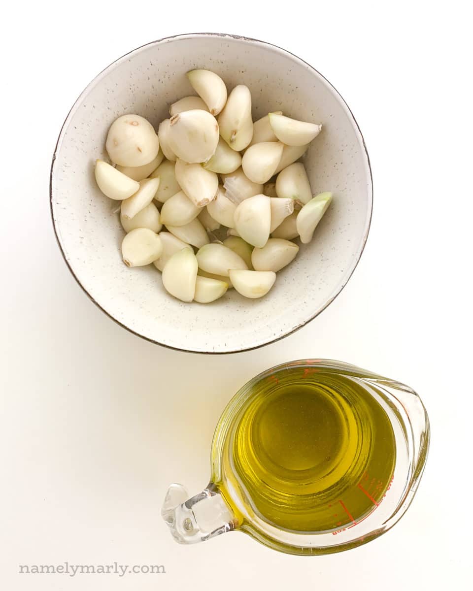 Peeled garlic is in a bowl next to a measuring cup full of olive oil.