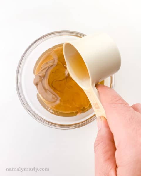 A hand holds a measuring cup full of syrup and is pouring it over a glass bowl full of peanut butter.