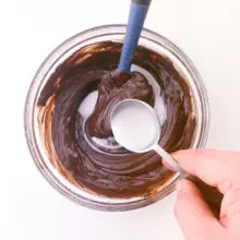 A hand holds a spoon pouring milk into a bowl with melted chocolate. There is a blue spatula in the bowl too.