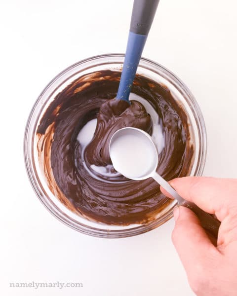 A hand holds a spoon pouring milk into a bowl with melted chocolate. There is a blue spatula in the bowl too.