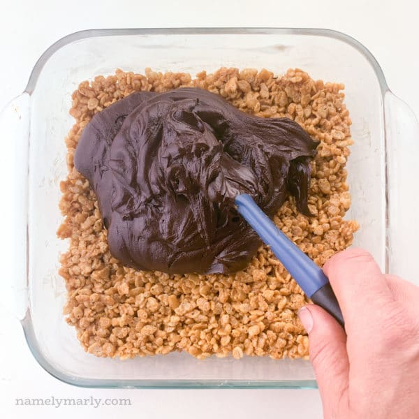 A hand holds a spatula and is spreading soft chocolate ganache over peanut butter rice crispies bars.
