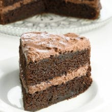 A thick slice of chocolate layer cake on a plate in front of the rest of the cake.