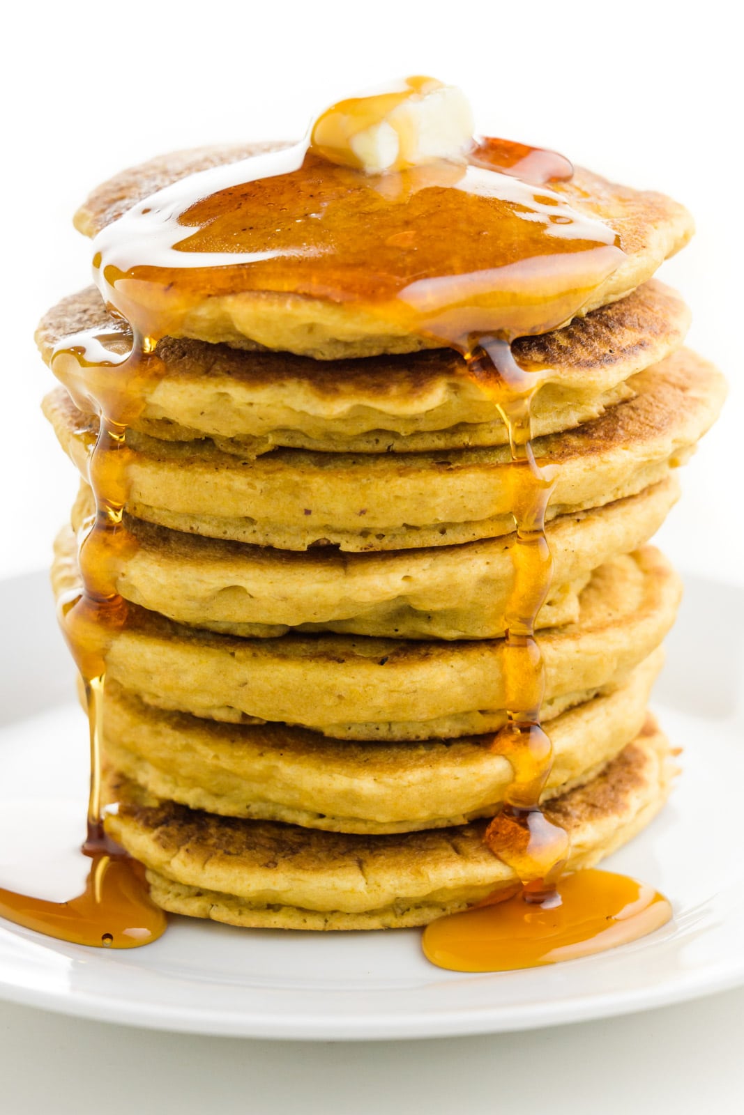 A stack of vegan pancakes has a pat of vegan butter on top and maple syrup flowing down the sides.