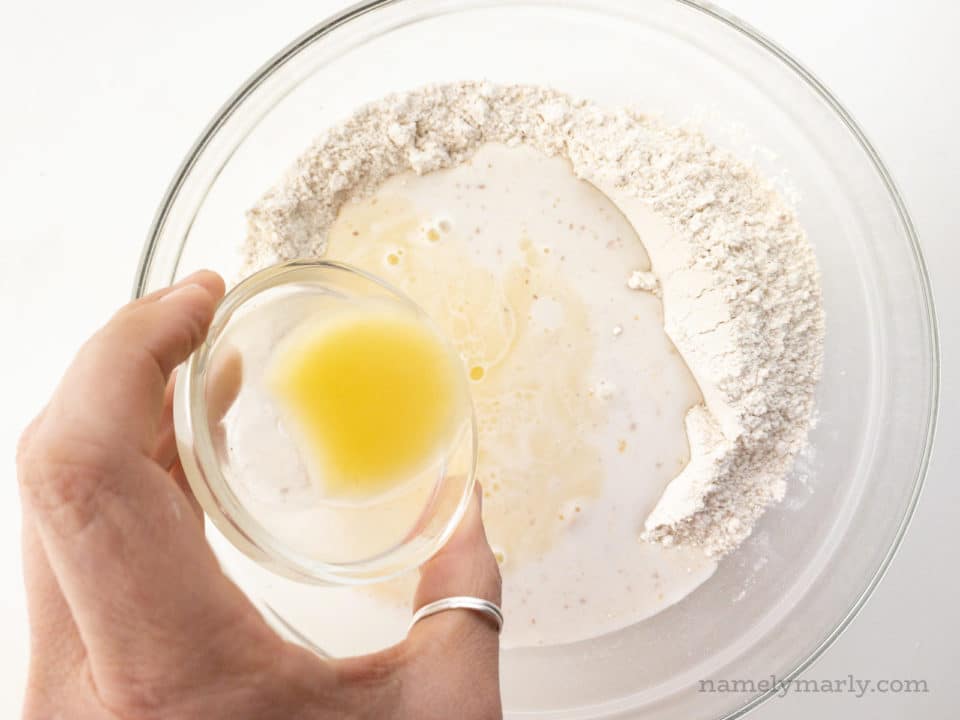 A hand holds a small glass bowl full of melted butter and is pouring it into a bowl with flour and almond milk.