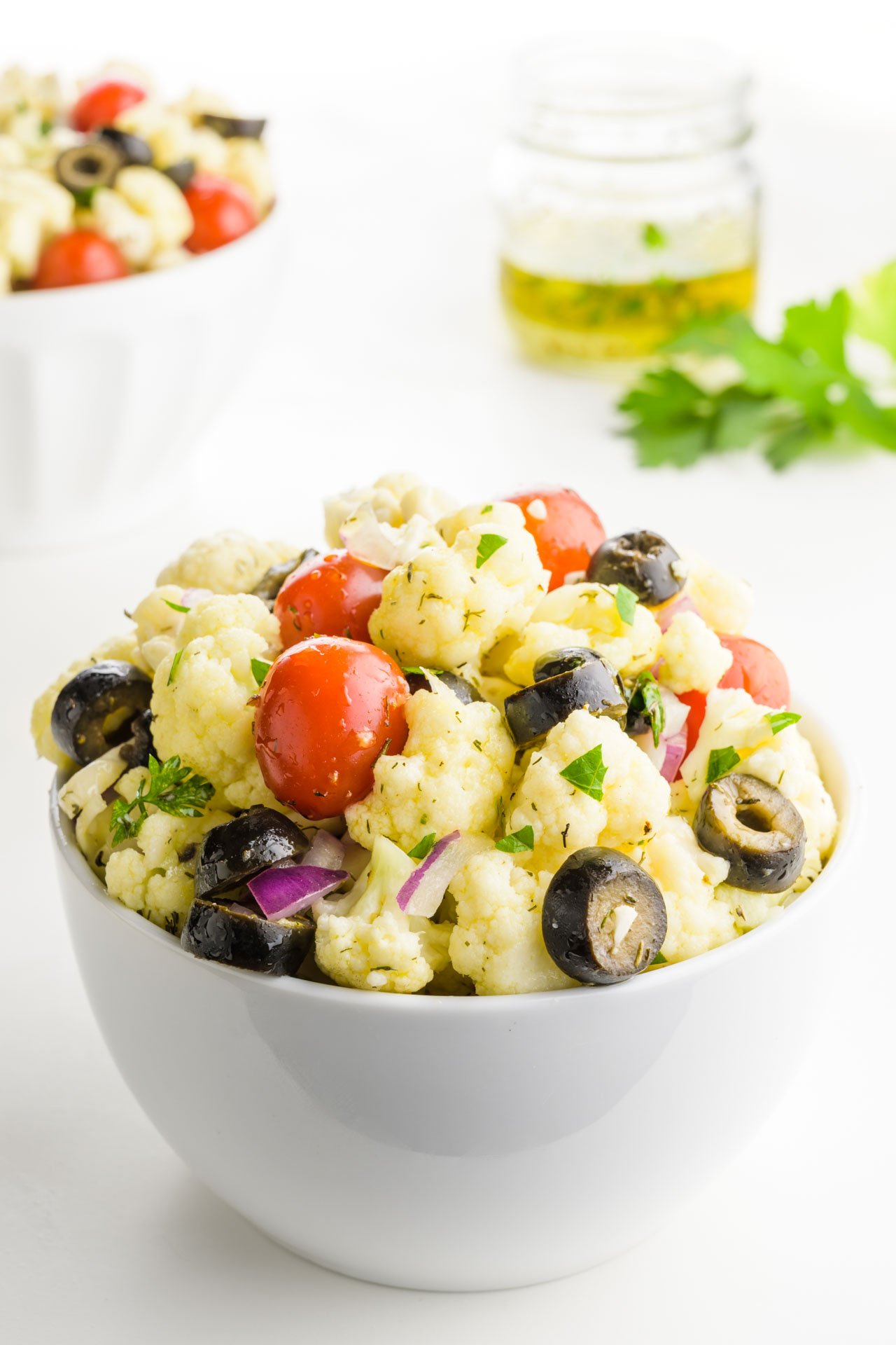 A bowl of cauliflower salad with another bowl of salad behind it and dressing and parsley behind it as well.