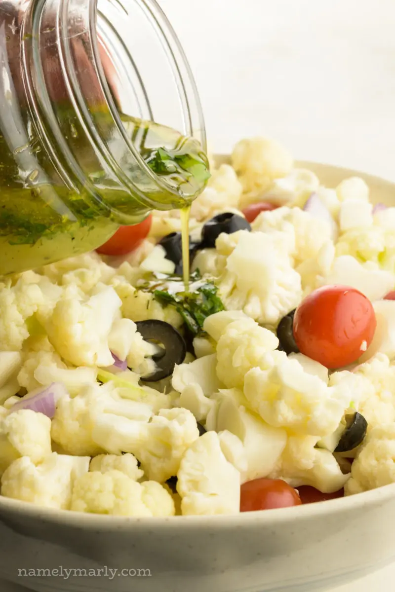 A small bowl of dressing is being poured over a bowl full of cauliflower florets, cherry tomatoes, and other veggies.
