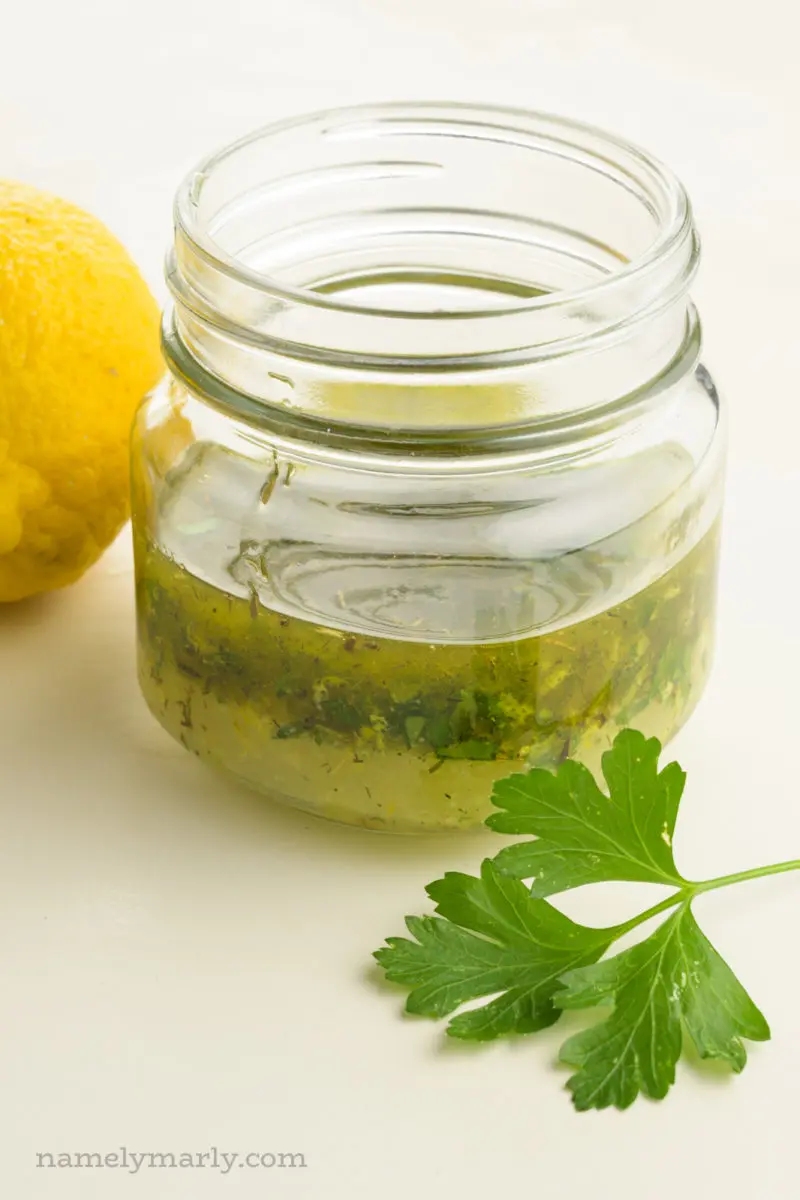 A small glass jar holds dressing, with a lemon behind it and a parsley leaf in front of it.