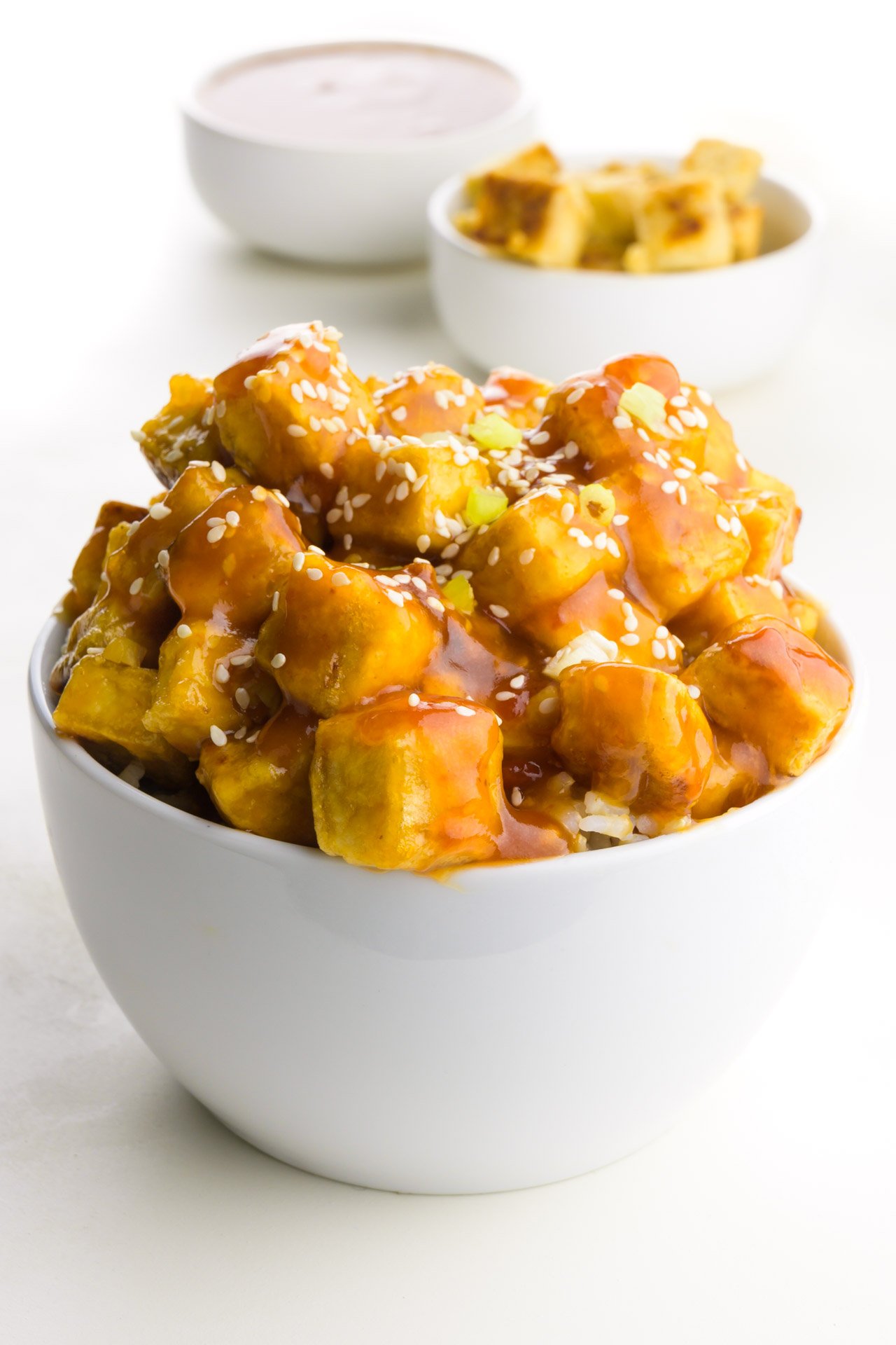 A bowl of tofu sits in front of a bowl of sauce and more tofu in the background.