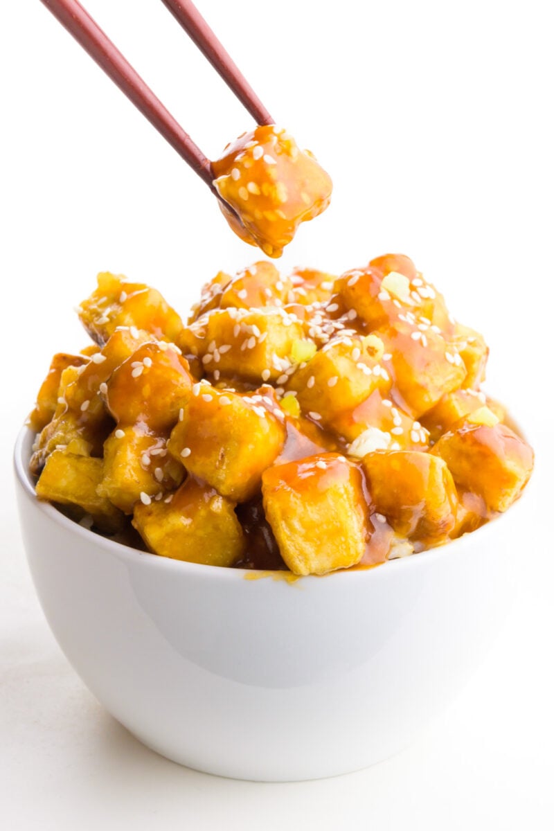 Chopsticks hold a single bite over a bowl full of sesame tofu.