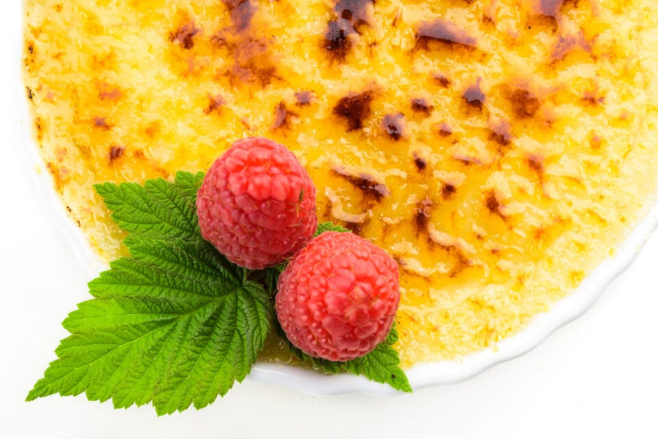A close-up shot of creme br&ucirc;l&eacute;e in a ramekin with burnt sugar on top and raspberries on the side.