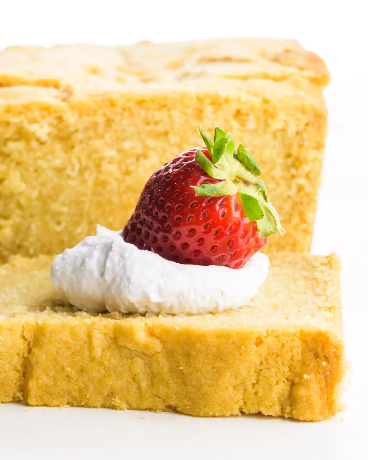 A slice of vegan pound cake has whipped cream and a strawberry on it. The rest of the pound cake is behind it.