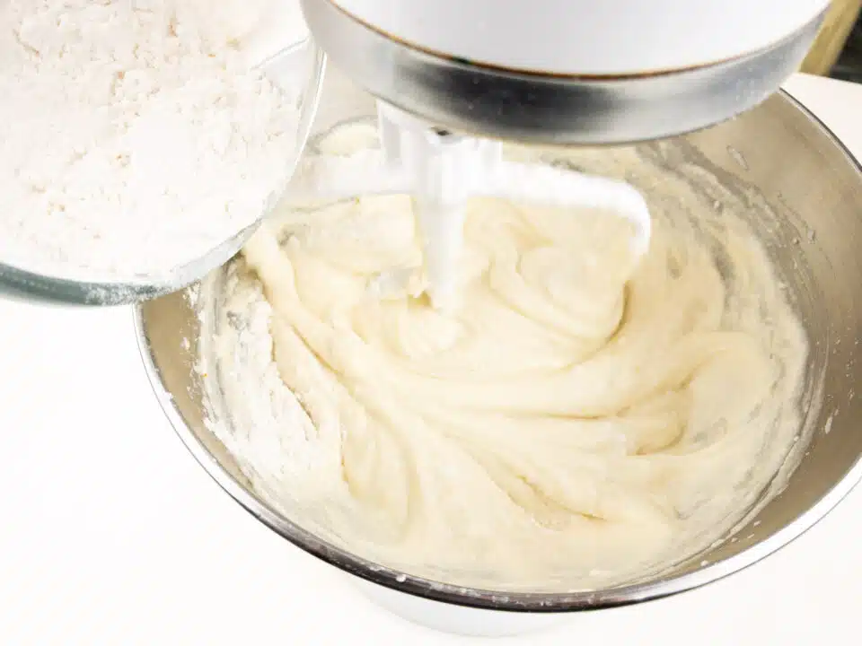 Flour is being poured into a stand mixer with a creamy batter.