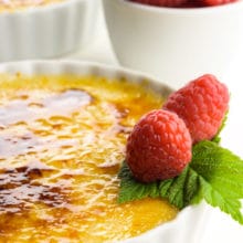 A close up of creme brûlée in white serving dishes with raspberries on top and a bowl of raspberries beside them.