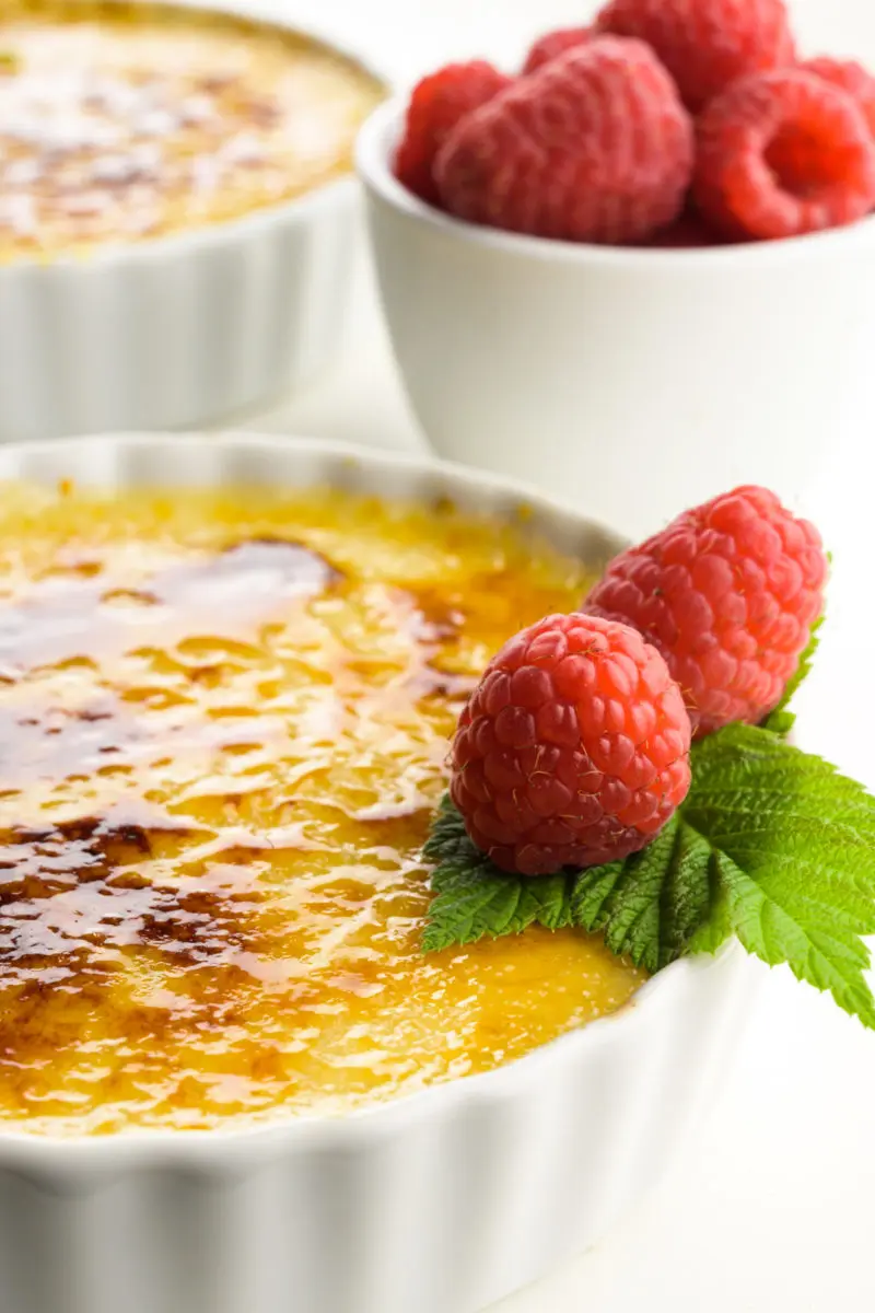 A close up of creme brûlée in white serving dishes with raspberries on top and a bowl of raspberries beside them.
