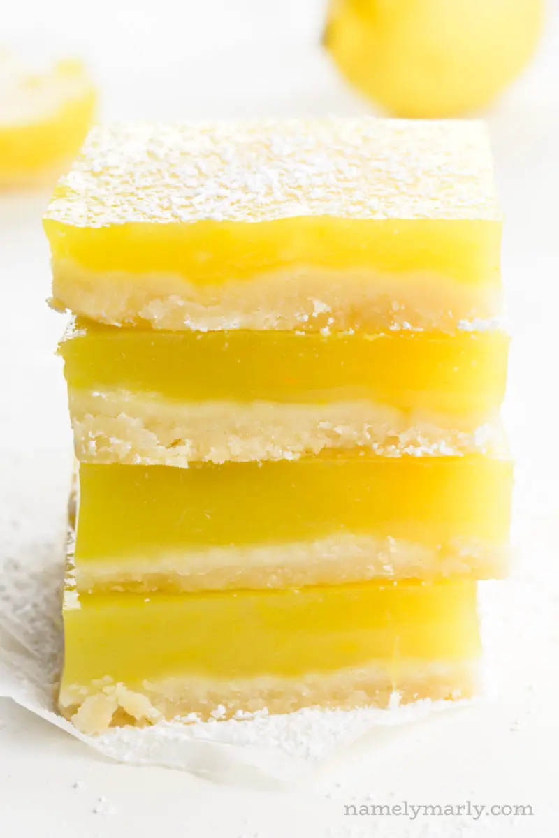 A stack of four lemon squares with more bars and a lemon behind it.