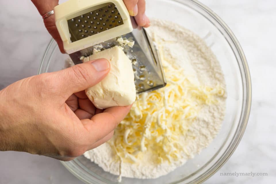 A hand holds butter, grating it over a bowl of flour mixture.