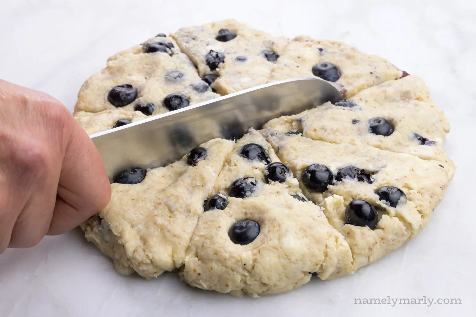 Dough with blueberries is in a disk and hand holding a knife is cutting it into slices.