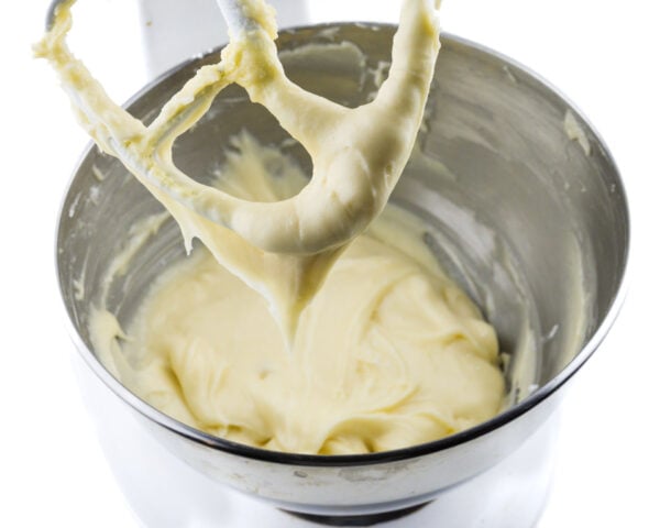 A stand mixer mixing bowl holds creamy frosting, with some of it on the beater.