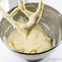 A stand mixer mixing bowl holds creamy frosting, with some of it on the beater.