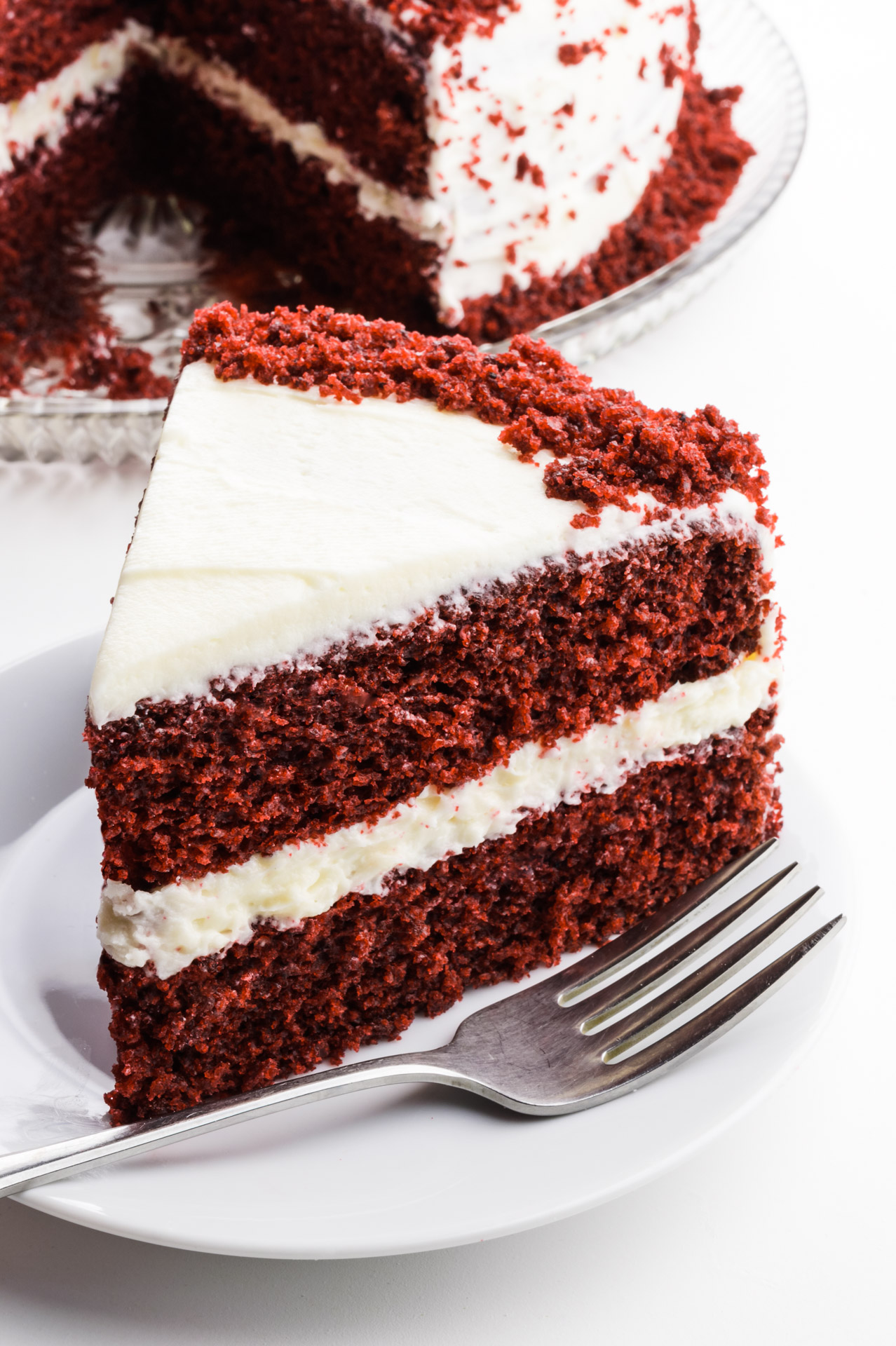 A slice of layered cake on a plate has a fork beside it and sits in front of the rest of the cake.