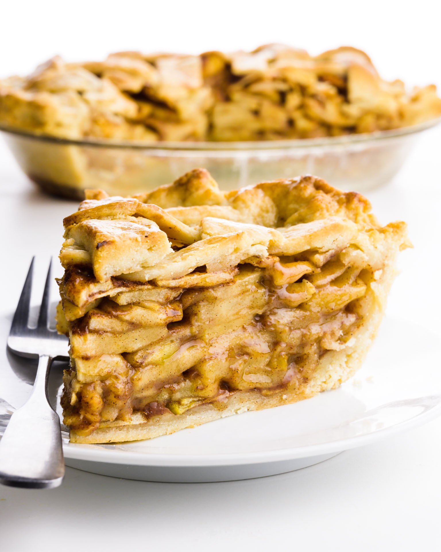 A slice of apple pie has a fork next to it and it sits in front of the rest of the pie.