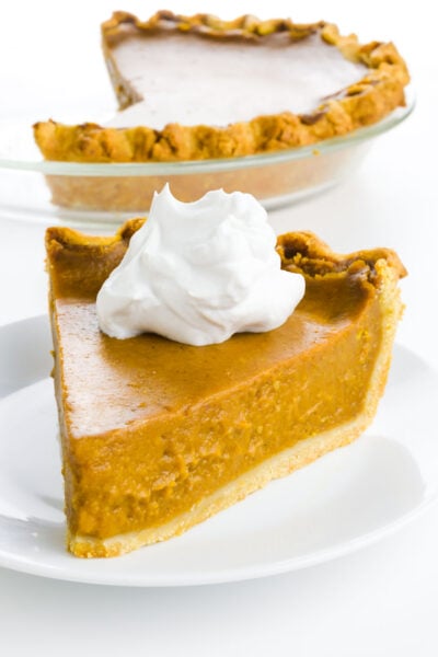 A slice of vegan pumpkin pie with a dollop of whipped cream sits in front of the rest of the pie.