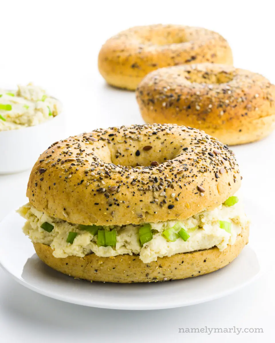 A bagel has been cut in half and has cashew cream cheese in the middle. There are more bagels behind it and a bowl of more vegan cream cheese.
