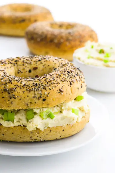 A bagel is cut in half and has vegan cream cheese in the middle. There are more bagels behind it with a bowl of more vegan cream cheese.