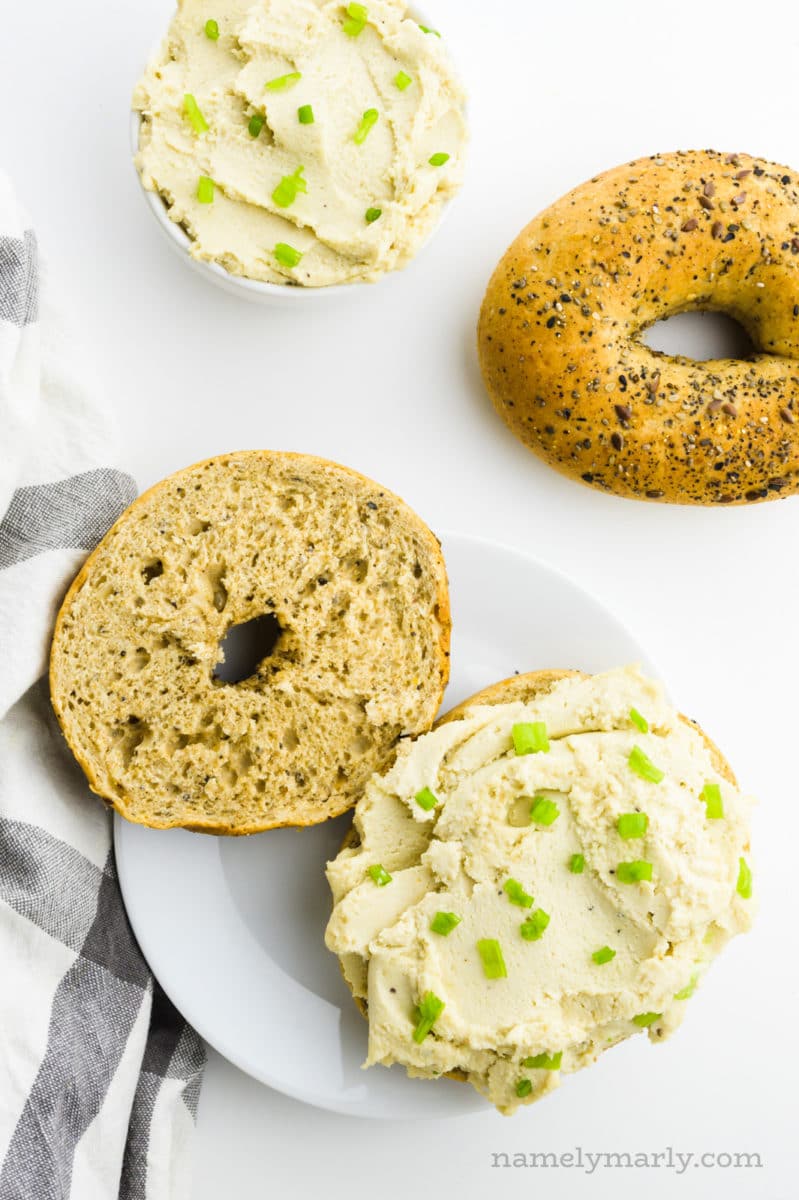 Looking down on a plate with a bagel cut in half. One half has vegan cream cheese over the top of it. There's another bagel behind the plate and a bowl with more cream cheese.