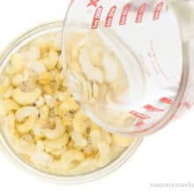 A bowl of raw cashews has been filled mostly with water. A pyrex measuring cup is finishing up pouring water over it.