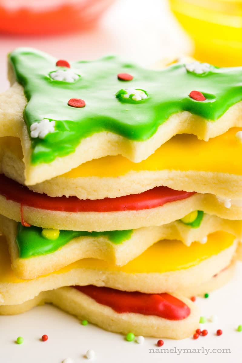 Several sugar cookies are stacked on top of each other with bowls of colorful frosting behind them.