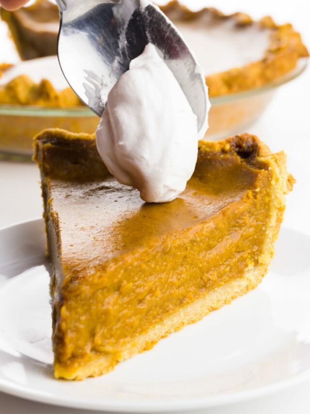 A hand holds a spoon dropping whipped cream on a slice of pumpkin pie.