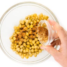 A hand pours spices over chickpeas in a bowl.