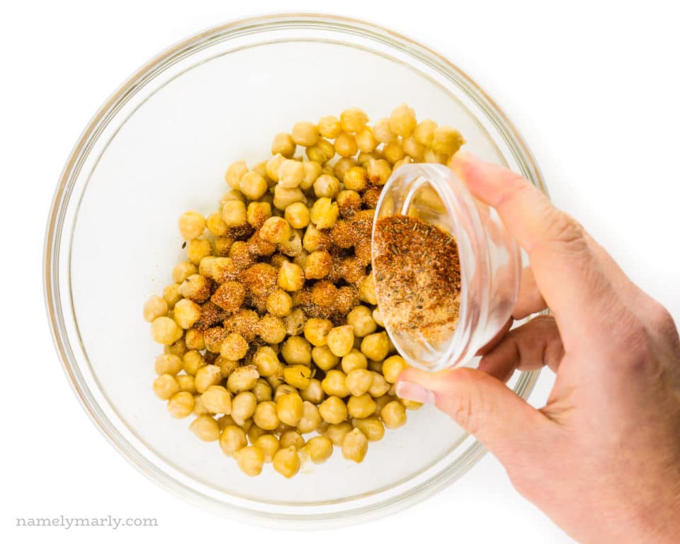 A hand pours spices over chickpeas in a bowl.