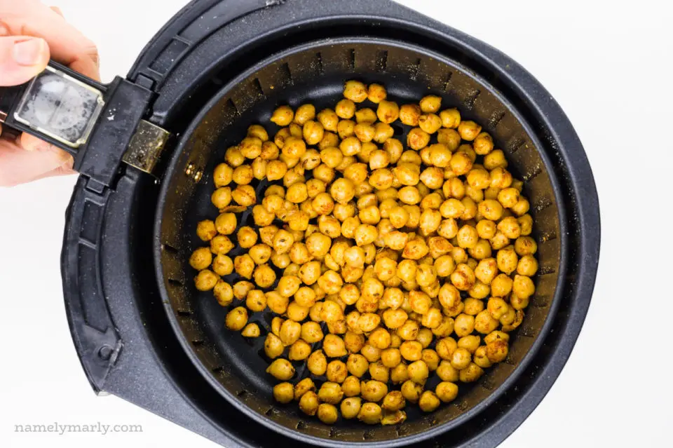 A hand holds an air fryer basket full of spiced chickpeas.
