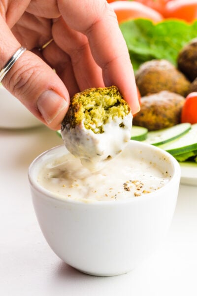 A hand holds an falafel and is dipping it in a white sauce.
